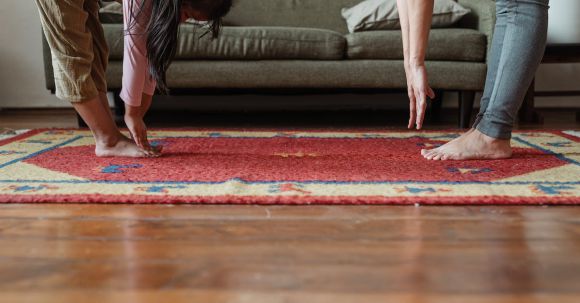 Floor - Side view of young ethnic woman stretching body with adorable girl while standing against each other on floor carpet near cozy sofa in modern living room in morning